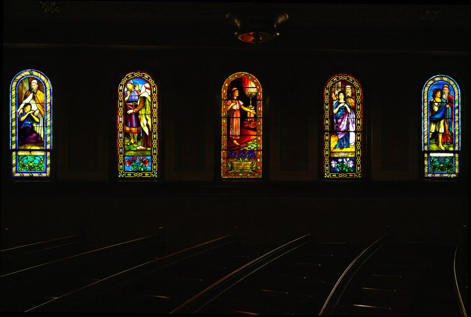 Four colorful stained glass windows depicting religious figures illuminate the dark interior of a church above empty pews.