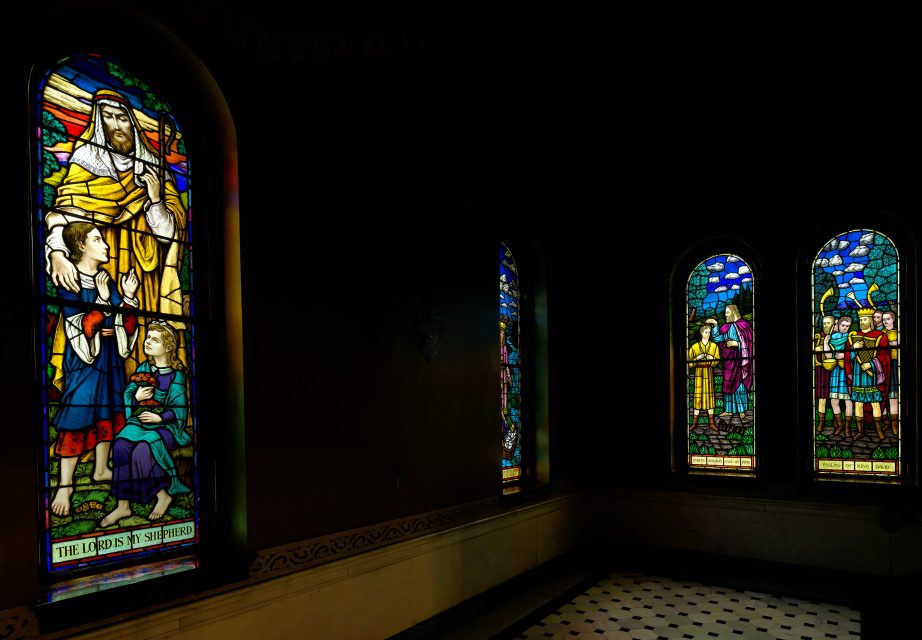 Dimly lit room with three stained glass windows depicting biblical scenes; one includes children and an adult holding a staff, and two others show groups of figures.