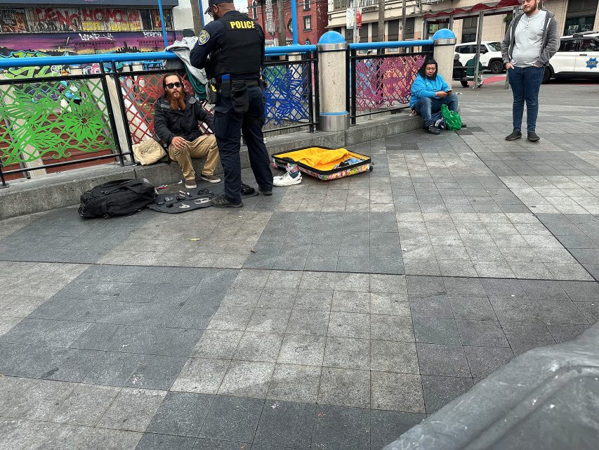 A police officer talks to a seated man with a guitar case on the ground; two other people sit nearby on a colorful fenced sidewalk in an urban area.