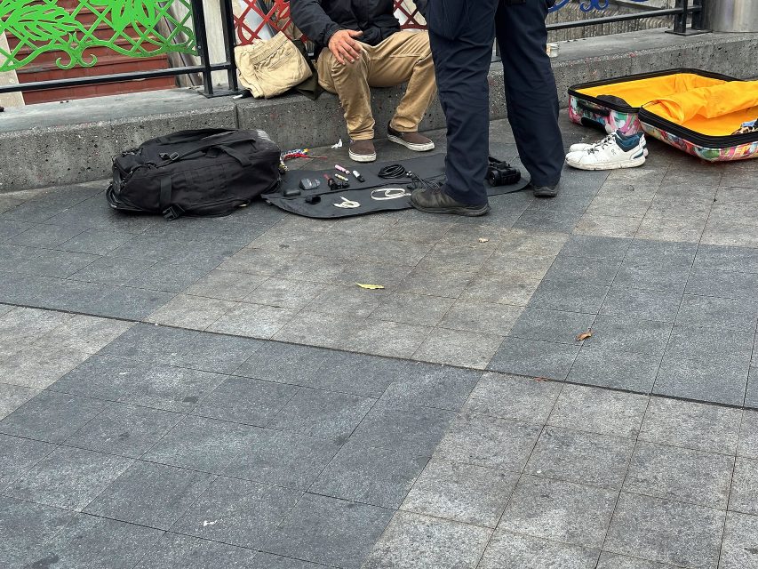 A person sitting on the pavement with belongings spread out, while another person in uniform stands nearby. An open suitcase and a backpack are on the ground.