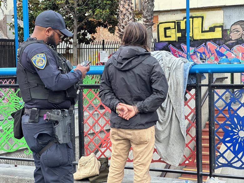 A police officer stands next to a person with hands behind their back near a fence covered with blankets and graffiti in an urban setting.