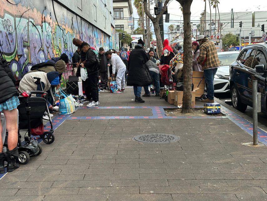 A group of people stand and sort through belongings on a city sidewalk near a wall with graffiti. Boxes, bags, and carts are visible on the ground.