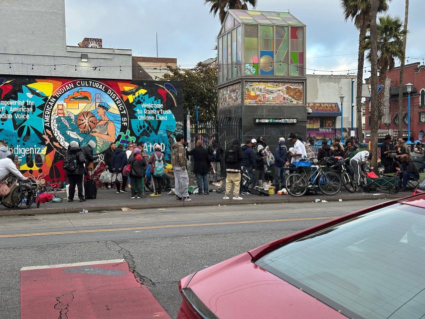 A large group of people gathers on a city sidewalk near a mural and a glass structure, with bicycles and belongings visible; a red car is in the foreground.