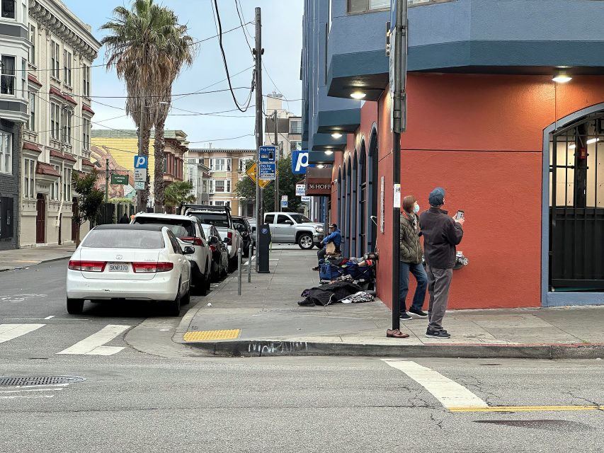 Two people stand on a sidewalk corner while another person sits with belongings near a red and blue building. Several parked cars and buildings line the street.