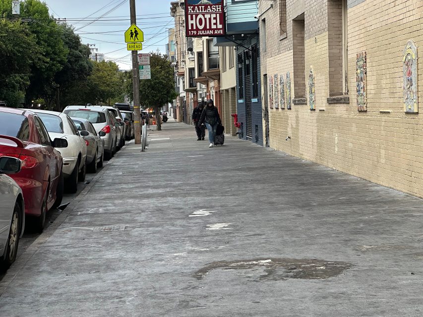 A sidewalk lined with parked cars beside a brick building with a sign reading "Kailash Hotel, 179 Julian Ave"; a few people walk in the distance.