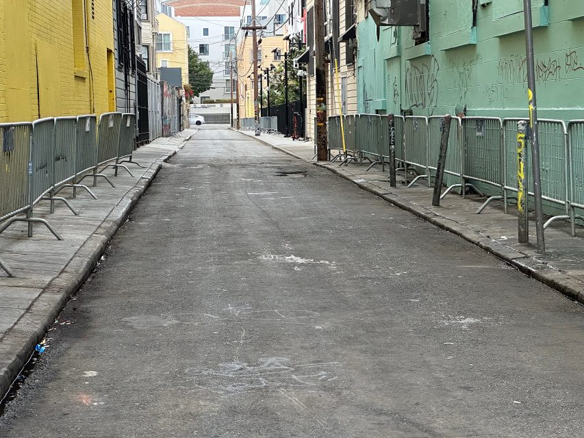 Narrow city alley lined with metal barricades on both sides, bordered by buildings with graffiti and faded paint.
