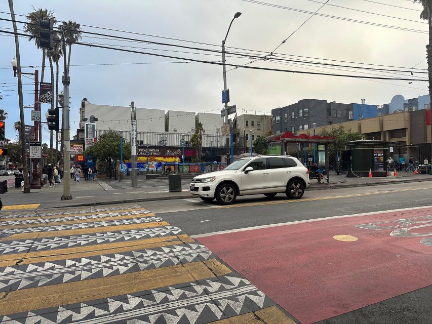 A white SUV waits at an intersection in an urban area with crosswalks, overhead wires, pedestrians, and surrounding buildings on a cloudy day.