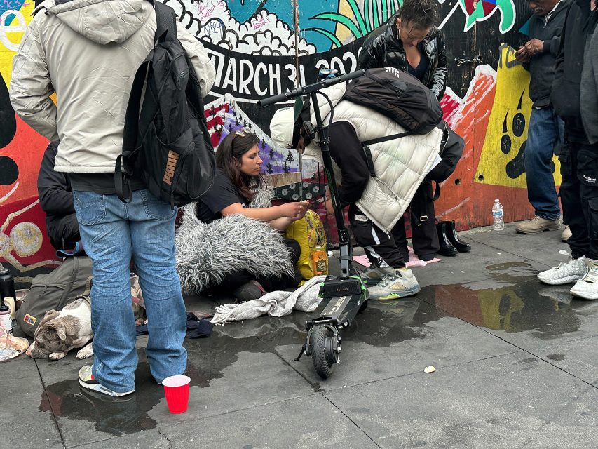Several people gather on a city sidewalk near a colorful mural, with belongings, a scooter, and a dog beside them. Puddles and personal items are scattered on the ground.