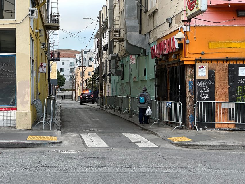 A narrow urban alleyway with metal barricades, a person walking, a parked car, and buildings with graffiti and boarded-up storefronts.