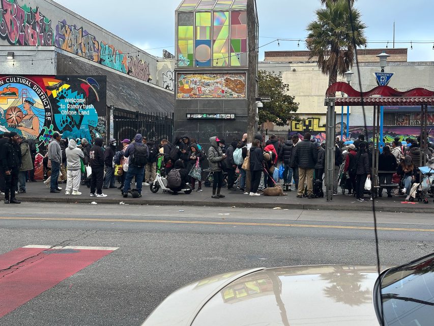 A group of people stand in line on a city sidewalk near colorful murals and a bus stop, with various bags and belongings visible.