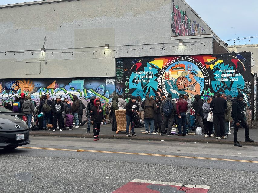 A crowd of people stands in line on a city sidewalk near a colorful mural that reads “Oakland Cultural District” and “Welcome to Ramaytush Ohlone Land.”.