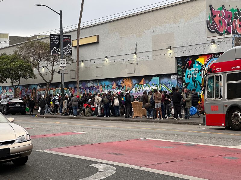 A crowd of people stands in line on a city sidewalk next to a building with colorful graffiti. A public bus and several cars are nearby.