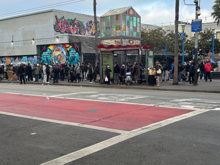 A long line of people waits outside a colorful mural-covered building near a glass bus shelter on an urban street.