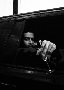 A person sitting in a car holds a wine glass out of the window, looking forward with a neutral expression. The image is in black and white.