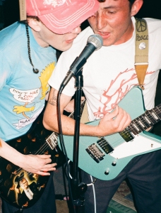 Two musicians playing electric guitars and singing into a shared microphone during a live performance.
