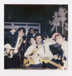 Five people are posing indoors, some holding electric guitars. They are smiling and making peace signs, with decorative white feathers visible in the background.