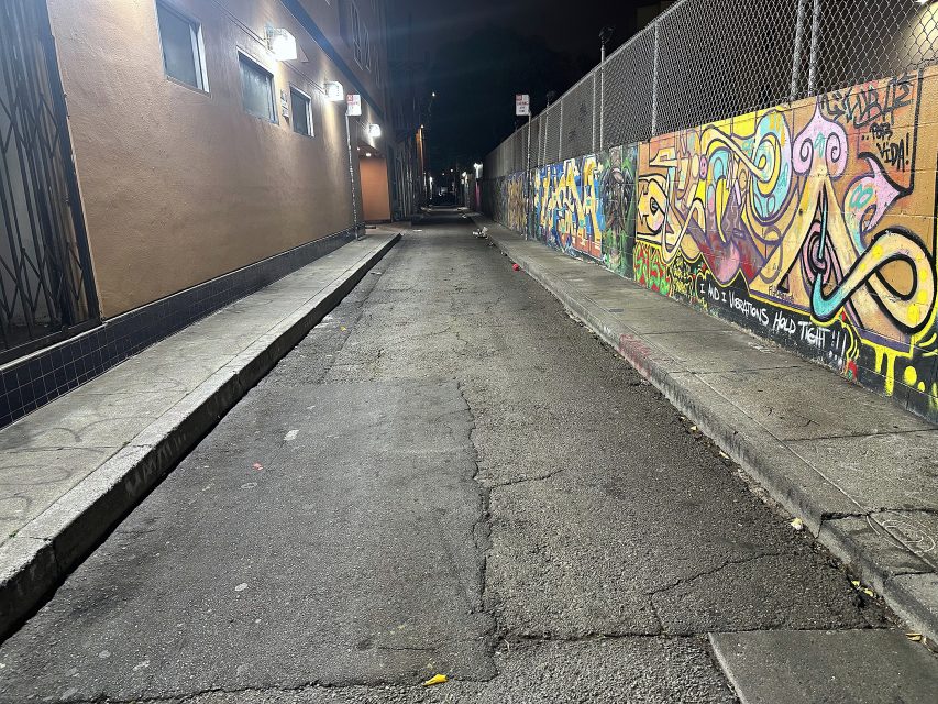 A narrow, empty alley at night is lit by overhead lights, with graffiti-covered walls on the right and a building with barred windows on the left.