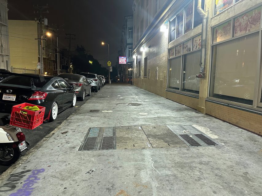 A dimly lit city sidewalk at night with parked cars on the left and a building with illuminated windows and signs on the right.