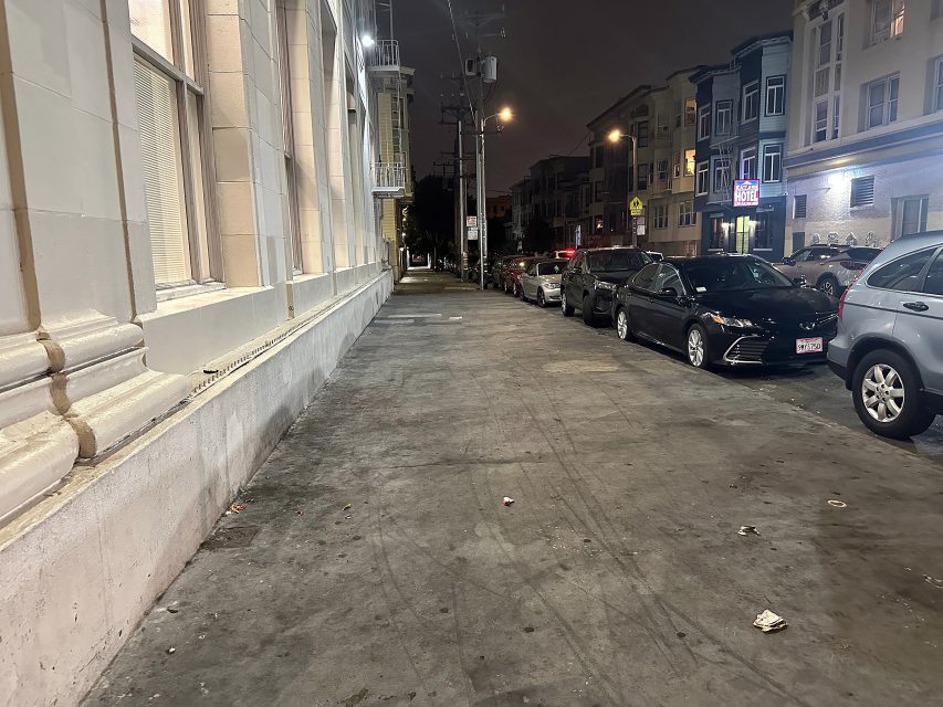 A city sidewalk at night with cars parked along the street and apartment buildings in the background. The sidewalk is mostly empty, with some litter scattered on the ground.