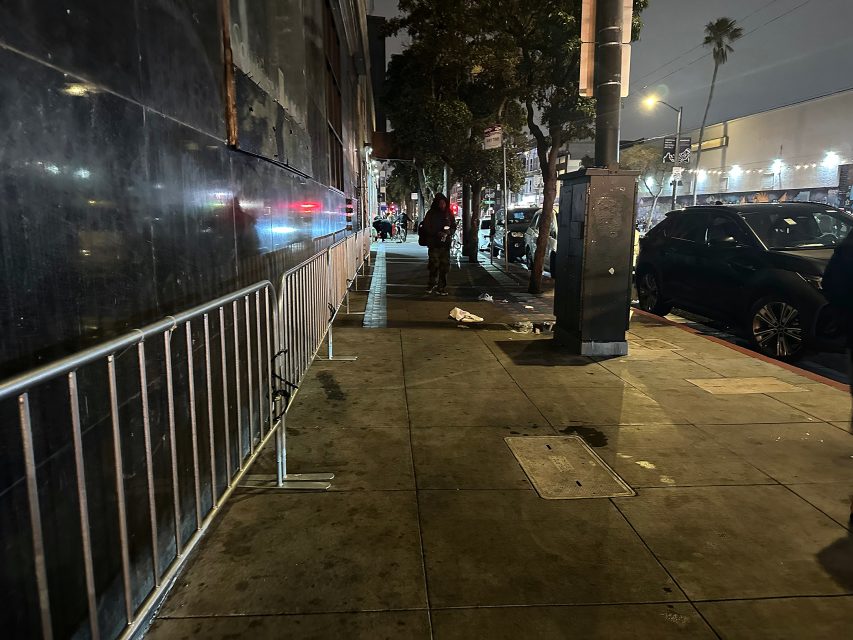 A dimly lit city sidewalk at night with metal barricades, a few pedestrians, parked cars, and streetlights in the background.