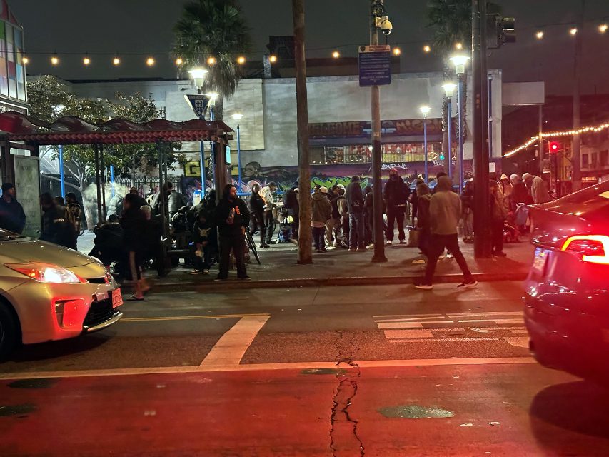 A crowd of people gathers on a city sidewalk at night, illuminated by streetlights and surrounded by parked cars and street art.