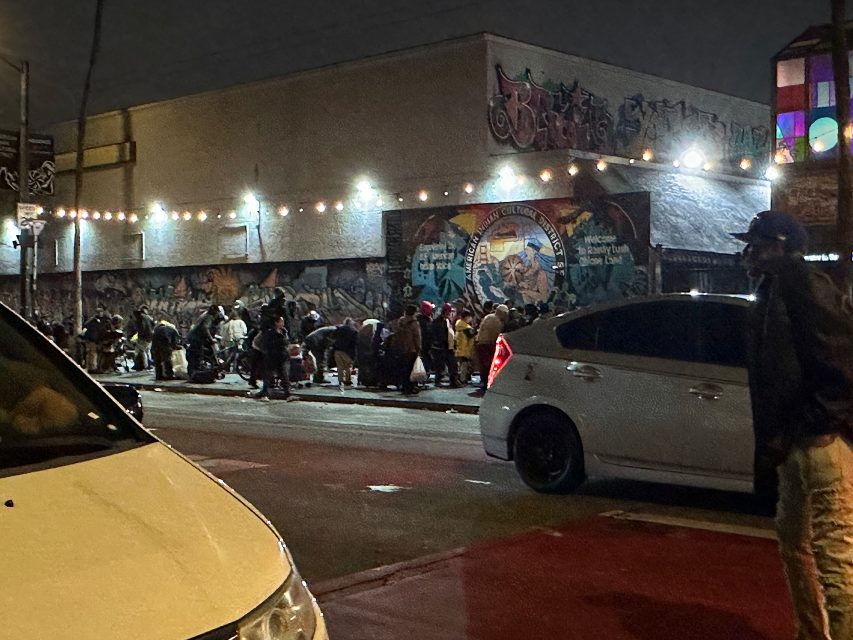 A group of people gathers on a city sidewalk at night near a mural and parked cars, with string lights hanging overhead.