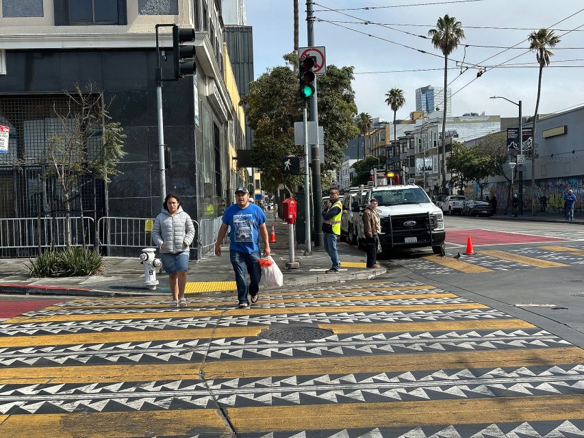 People cross a city street at a crosswalk with bold geometric patterns. A truck, workers in safety vests, and traffic lights are visible. Buildings and palm trees line the background.