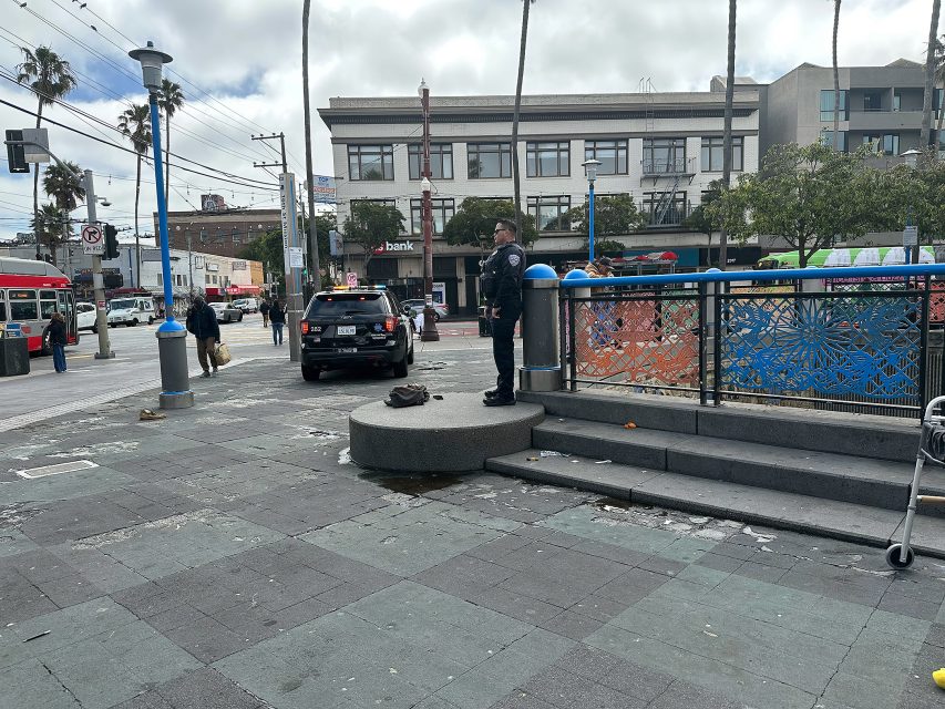 A police officer stands on a small platform near steps in an urban plaza beside a parked patrol car, with people and buildings in the background.
