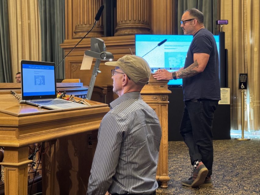 Two men present at a lectern in a formal room; one stands at a podium speaking while the other sits in front of a laptop, with presentation slides visible on nearby screens.
