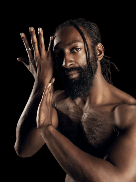 A shirtless man with a beard and dreadlocks poses with his hands raised and crossed in front of his face against a black background.