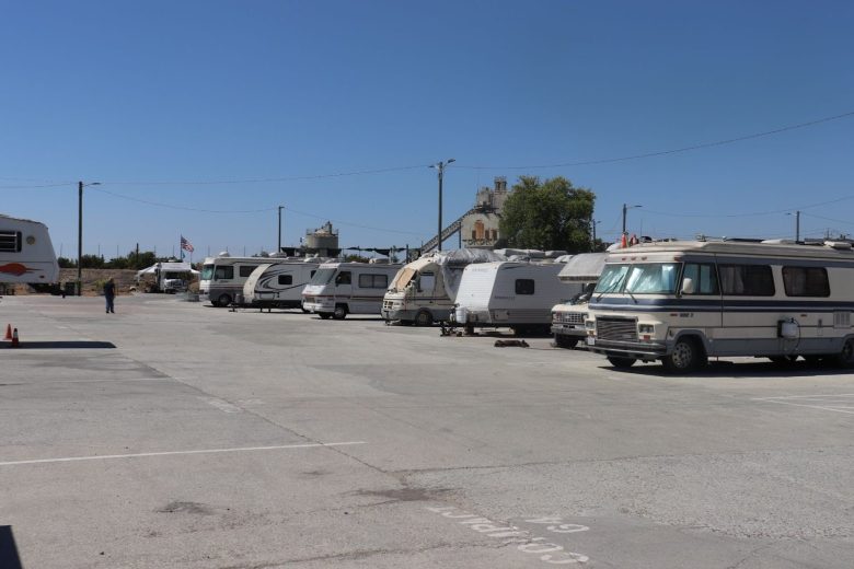 A row of RVs and trailers parked in a mostly empty, sunlit lot with a clear blue sky and a few buildings in the background.