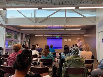 A group of people seated in a room face a blank blue projector screen as a man stands and speaks at the front.