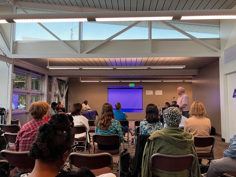 A group of people seated in a room face a blank blue projector screen as a man stands and speaks at the front.