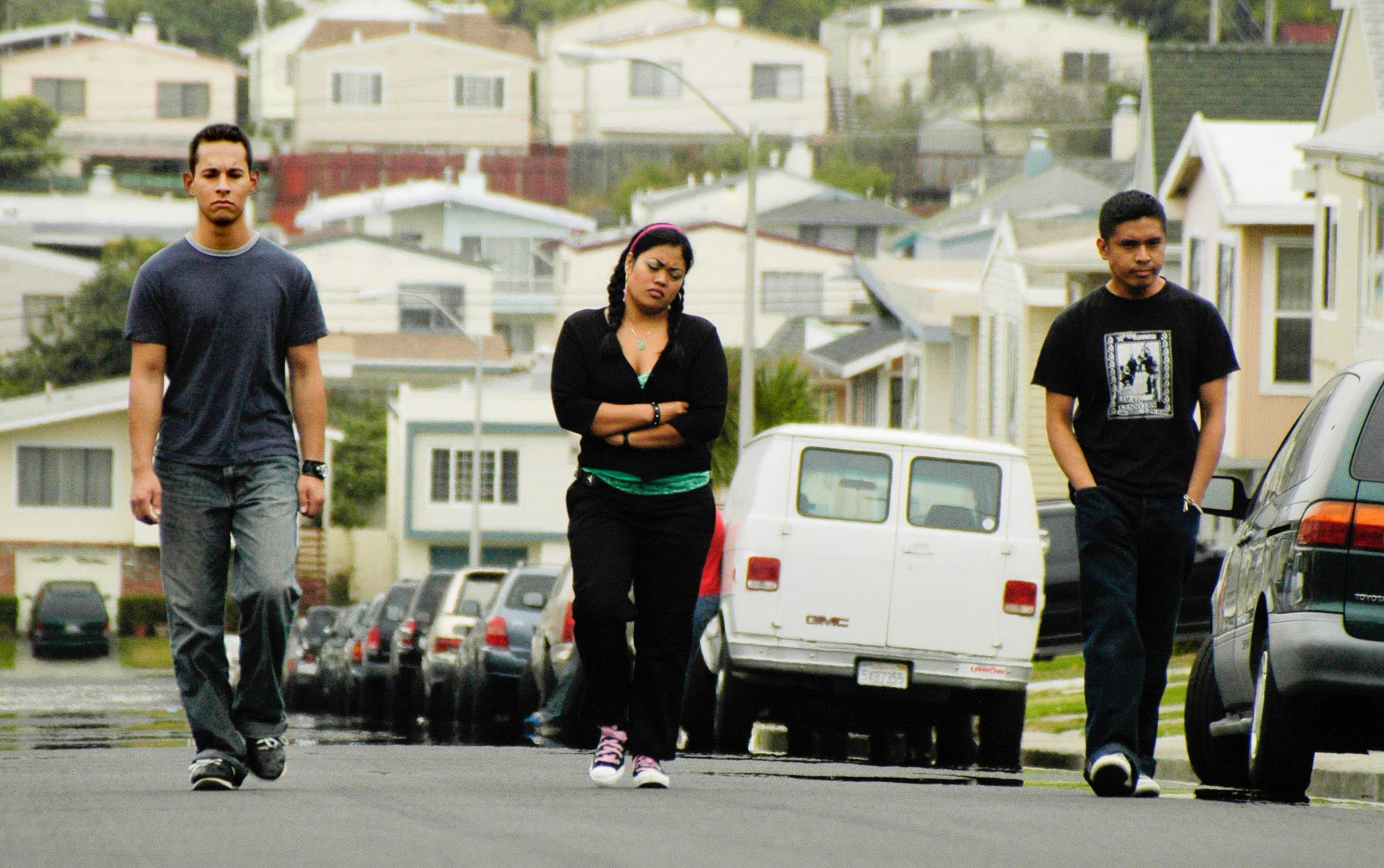 Three people walk separately down a suburban street lined with parked cars and houses on a cloudy day.