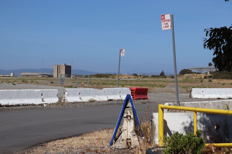 A fenced-off empty lot with concrete barriers, a "No Stopping Fire Lane" sign, and a distant building under a clear blue sky.