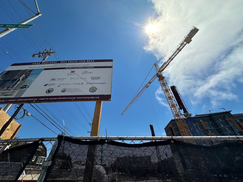 A construction site with a crane, a building under development, a sign displaying project information, and the sun shining in a clear blue sky.
