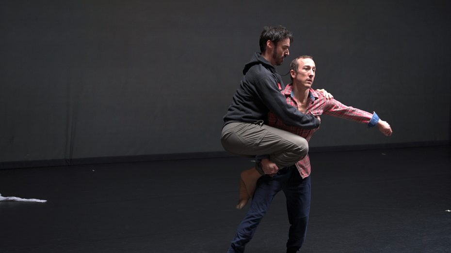 Two men perform a contemporary dance; one man in a hoodie is lifted off the ground, clinging to the other man in a plaid shirt on a plain stage.
