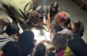 A group of people sit in a circle on the floor, closely examining and pointing at a large sheet of paper with drawings and notes.