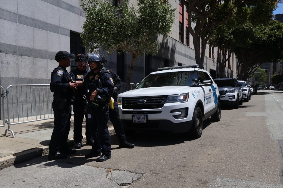Four police officers stand and talk next to parked police vehicles on a city street beside a large building during daylight.