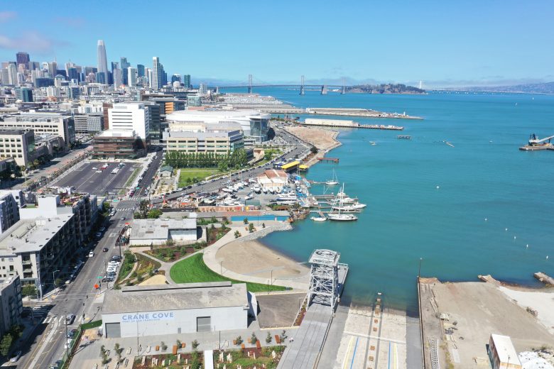 Aerial view of Crane Cove Park with docks, boats, and the San Francisco skyline in the background, next to the blue waters of the bay.