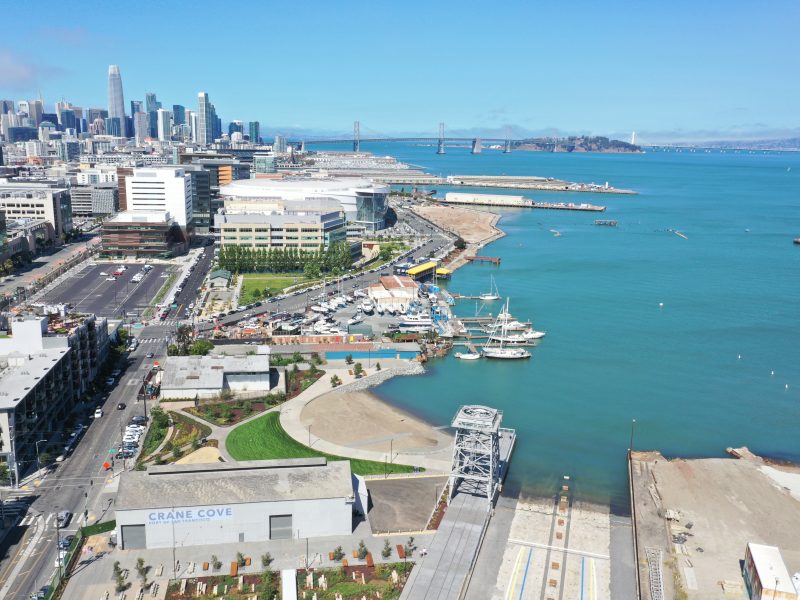 Aerial view of Crane Cove Park with docks, boats, and the San Francisco skyline in the background, next to the blue waters of the bay.
