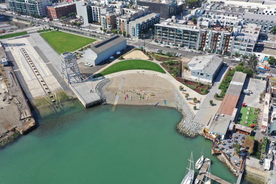 Aerial view of a waterfront park with green lawns, a dock, buildings, and a boat moored along the shoreline in an urban area.