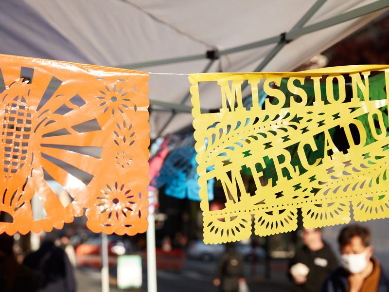 Colorful papel picado banners hang under a tent at an outdoor market. The yellow banner reads "Mission Mercado." People and city street are visible in the background.