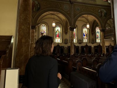 A person with short brown hair stands near the entrance of a church, facing stained glass windows and wooden pews inside a dimly lit interior.