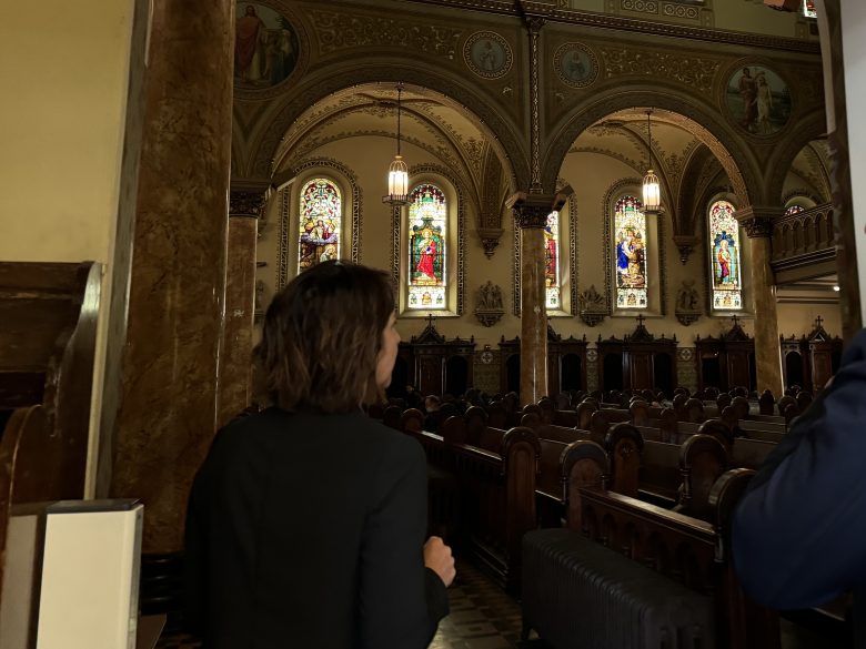 A person with short brown hair stands near the entrance of a church, facing stained glass windows and wooden pews inside a dimly lit interior.