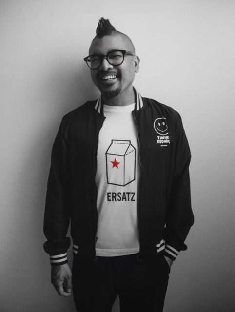 Smiling person with glasses and mohawk, wearing a black jacket and a white t-shirt with a milk carton graphic labeled โERSATZ.โ.
