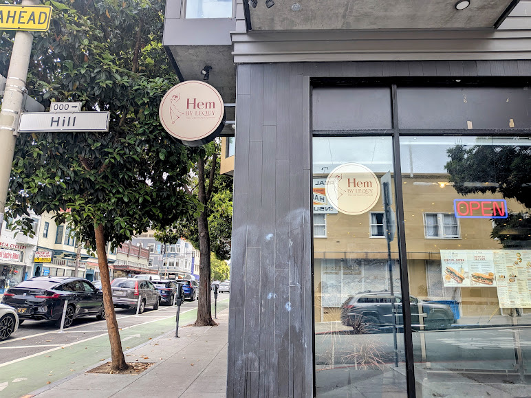 Street view of a corner restaurant with a sign reading "Hem By ELEGY," a visible menu in the window, and an illuminated "OPEN" sign. Cars are parked along the street.