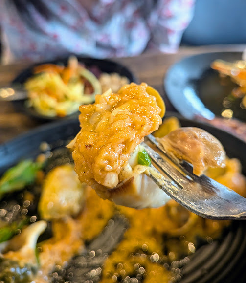 A close-up of a fork holding a bite of cheesy, saucy food with rice and vegetables, with a blurred plate of food and a person in the background.