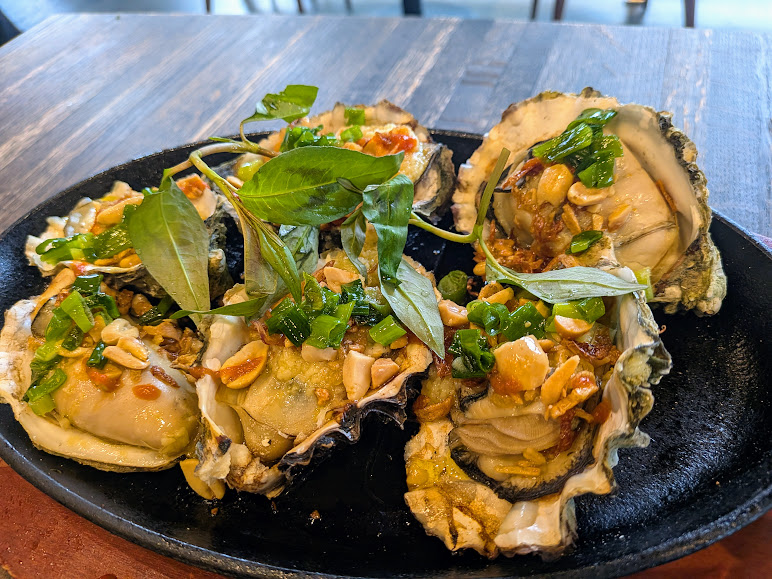 A plate of cooked oysters topped with chopped peanuts, green onions, herbs, and sauce, served on a black dish.