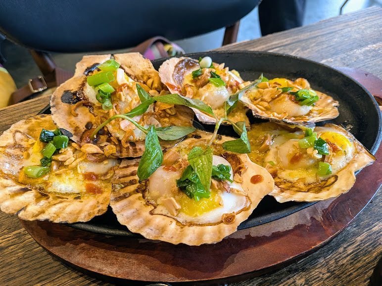 Grilled scallops served on their shells, topped with chopped green onions, herbs, and crispy garlic, presented on a black plate over a wooden table.